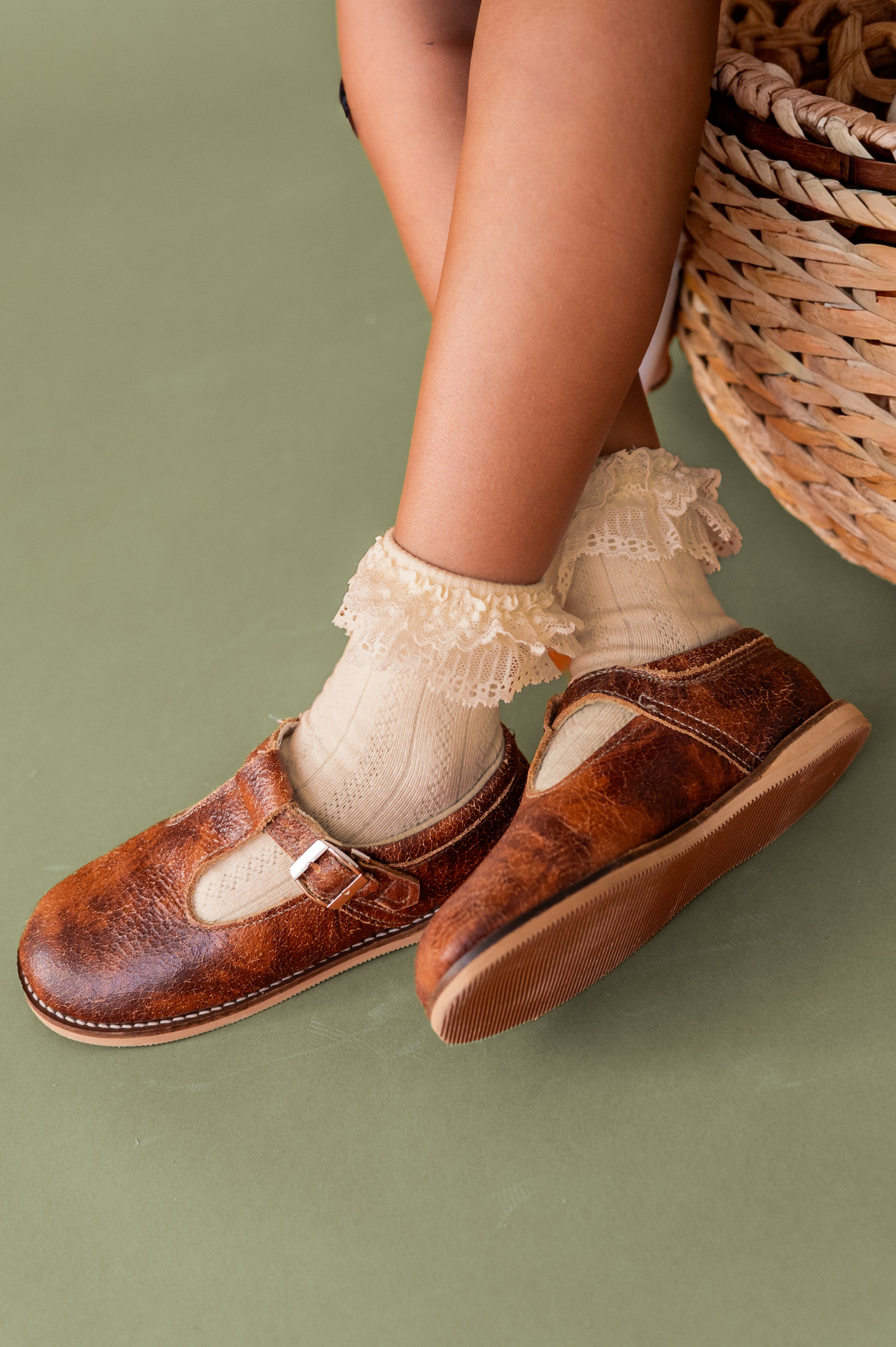 girls crossed feet in vintage cream lace top ankle socks and brown leather T-strap shoes on olive green background