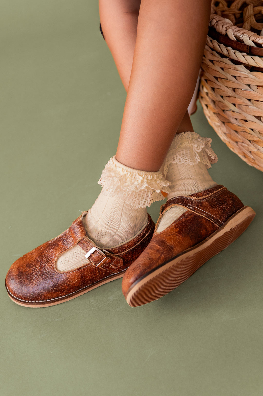 girls crossed feet in vintage cream lace top ankle socks and brown leather T-strap shoes on olive green background