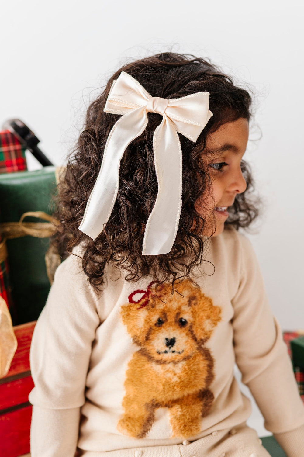 A young child wearing a cream knit sweater with a brown dog applique and a large cream bow in their hair, sitting next to wrapped Christmas presents.