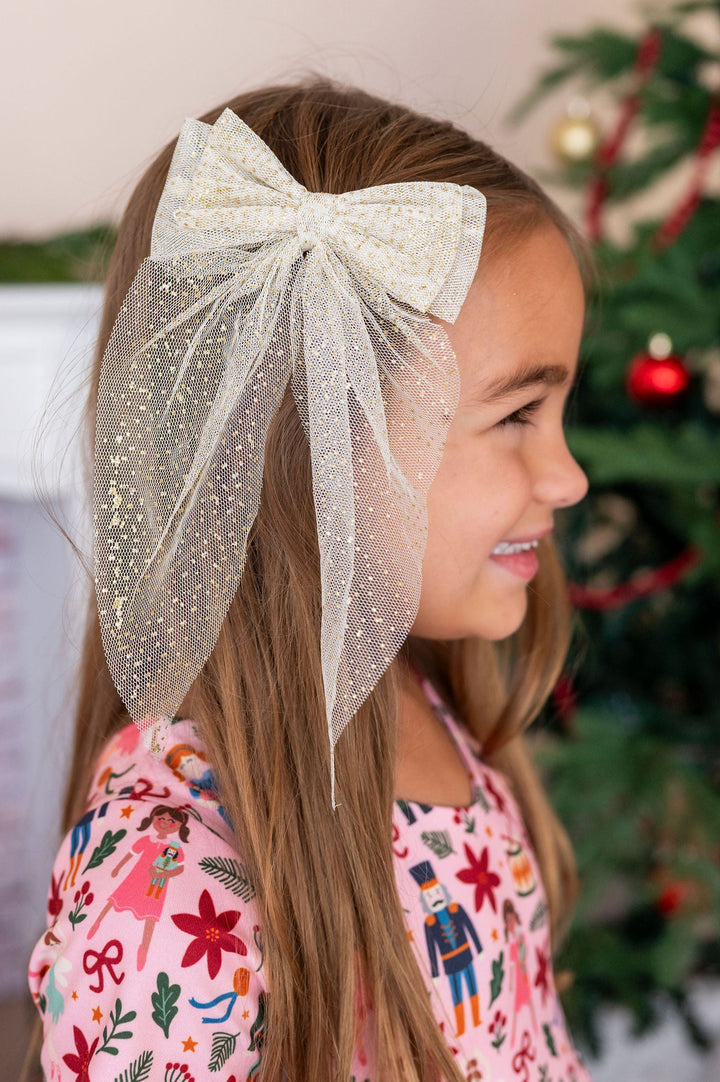 side profile of little girl wearing pink nutcracker inspired dress with large cream  gold sparkle tulle hair bow with a blurred holiday backgroundnd