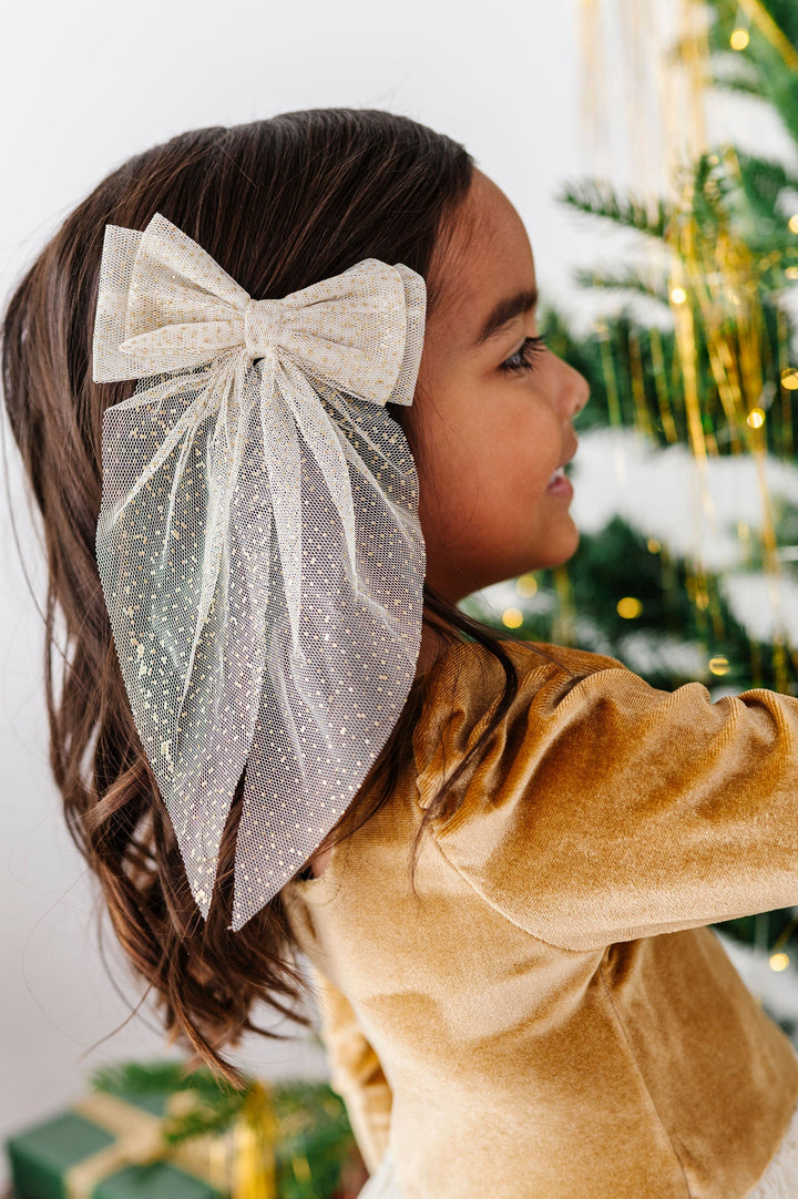 little girl wearing golden velvet dress decorating xmas tree wearing a large vanilla and gold sparkle tulle hair bow