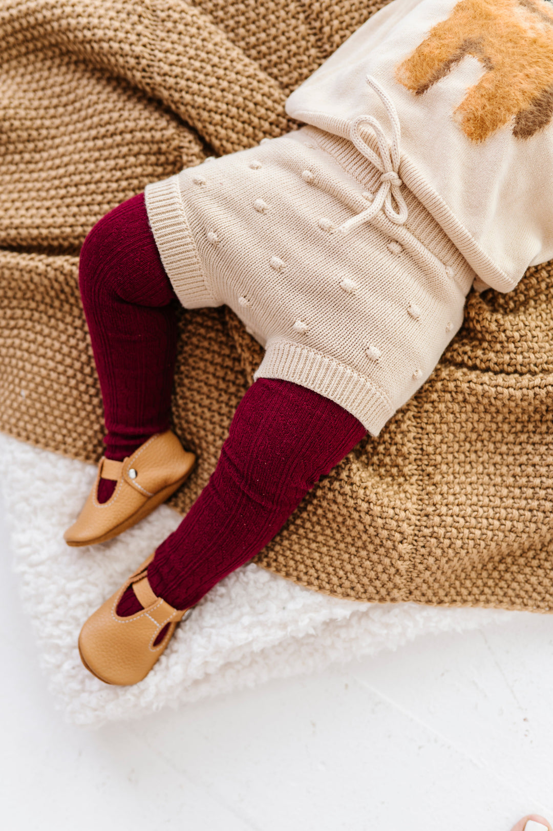 baby girl on blanket in cream knit bloomers with faux tie at the waist and 3D bubble texture paired with cream puppy sweater and burgundy cable knit tights