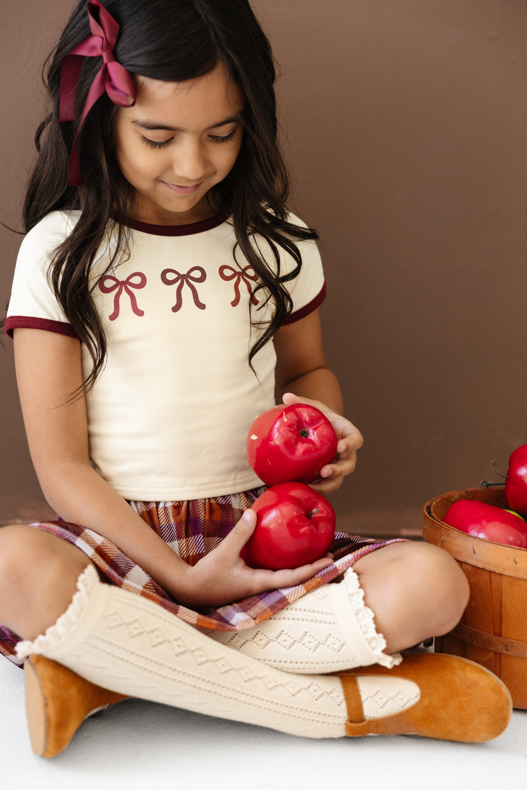 little girl holding apples in cream top with burgundy binding and bow print paired with matching burgundy satin hair bow, fall plaid velvet skort and cream open knit knee highs with ruffle