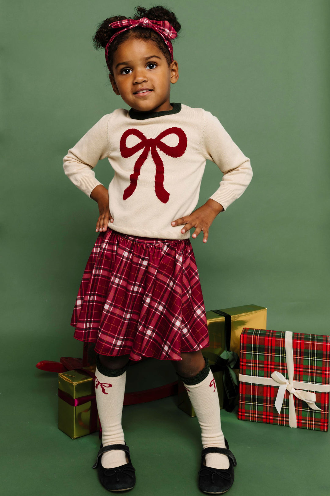 little girl with Christmas gifts in matching cream sweater and knee socks with crimson bow design paired with crimson plaid skirt and headband 