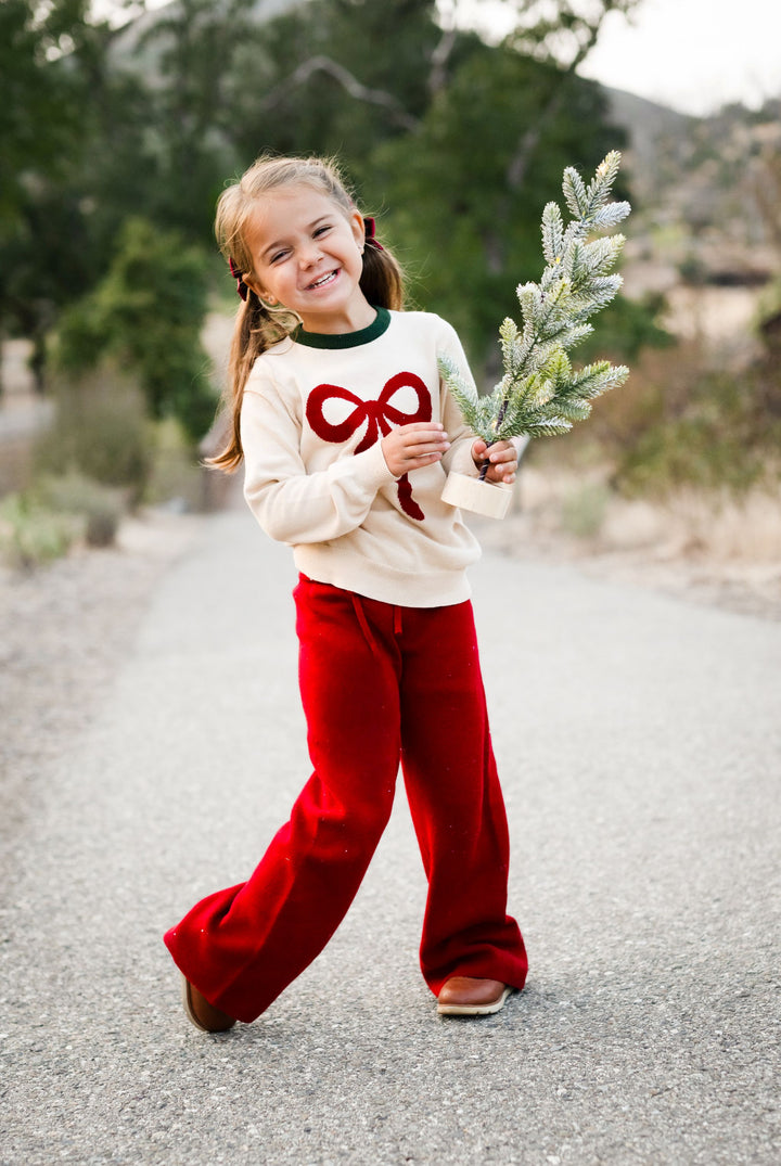 little girl in forest holding small Christmas tree in cream cotton knit sweater with red bow design and matching red sweater pants