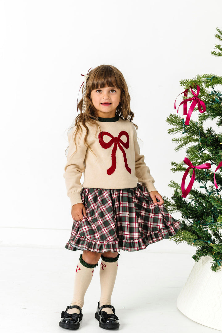 little girl by Christmas tree in green plaid twirl dress, cream sweater with crimson bow and forest green neck nd matching bow knee socks
