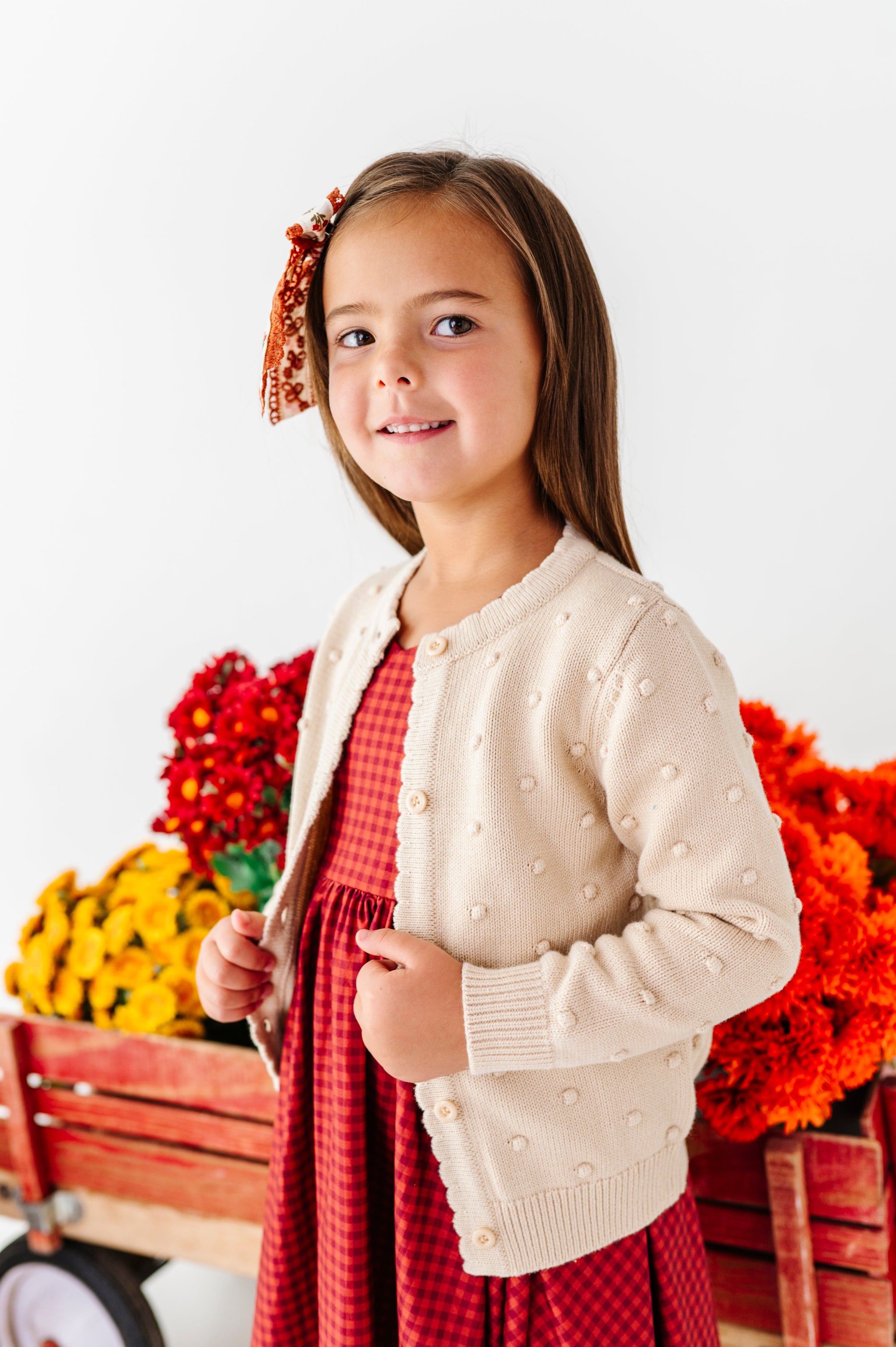 little girl in front of fall flower filled wagon in cream bubble knit cardigan and rust gingham twirl dress