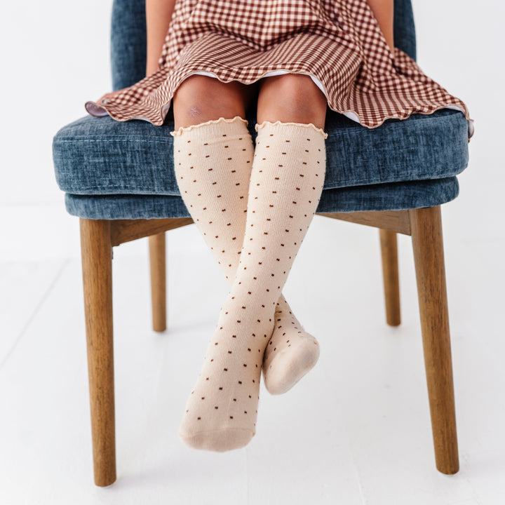 little girl sitting on blue chair in cream lettuce trin knee socks with tiny brown dots paired with brown gingham fall dress