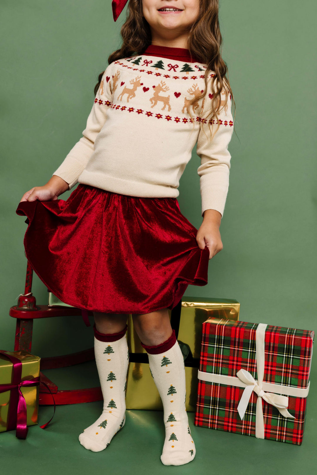 little girl by Christmas gifts in cream knit sweater with fair isle reindeer design, matching socks and crimson velvet skirt