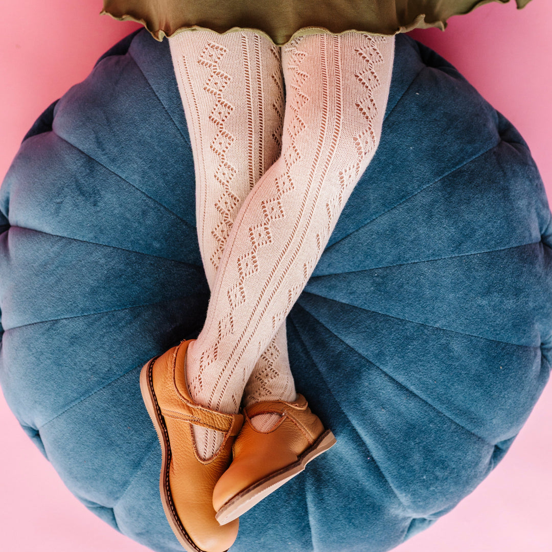 young girl sitting on teal velvet pillow wearing cream open knit tights with brown mary janes and olive twirl dress