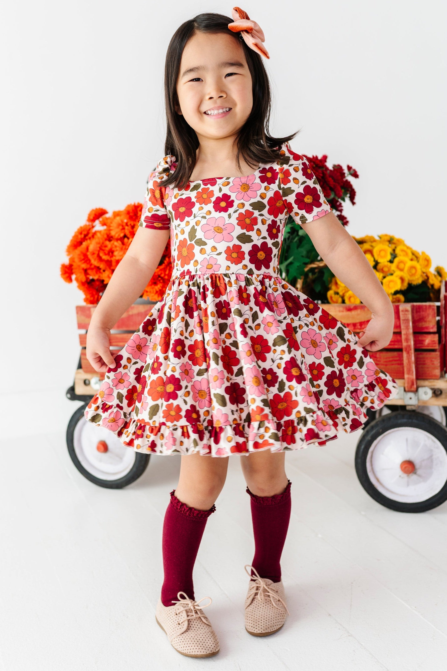 young girl in front of fall flower wagon wearing cream fall floral print twirl dress, burgundy lace top knee socks and rust velvet hair bow