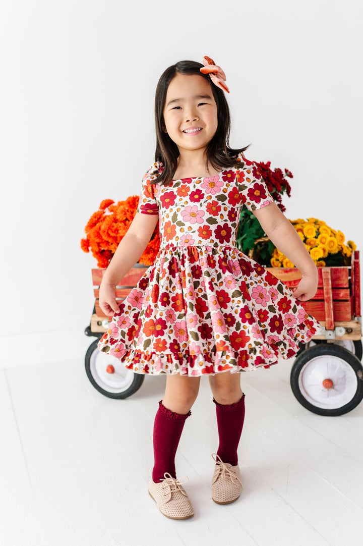 young girl in front of fall flower wagon wearing cream fall floral print twirl dress, burgundy lace top knee socks and rust velvet hair bow