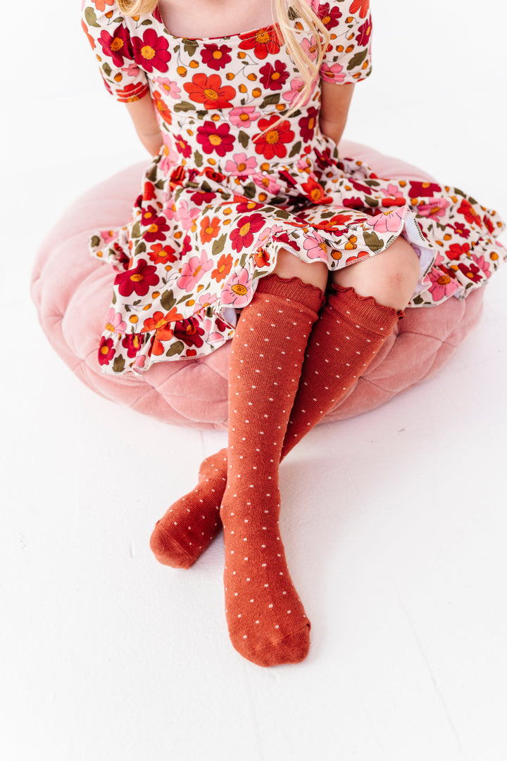 little girl sitting on pink velvet cushion in cream twirl dress with fall floral print paired with matching rust dotted knee socks with lettuce trim