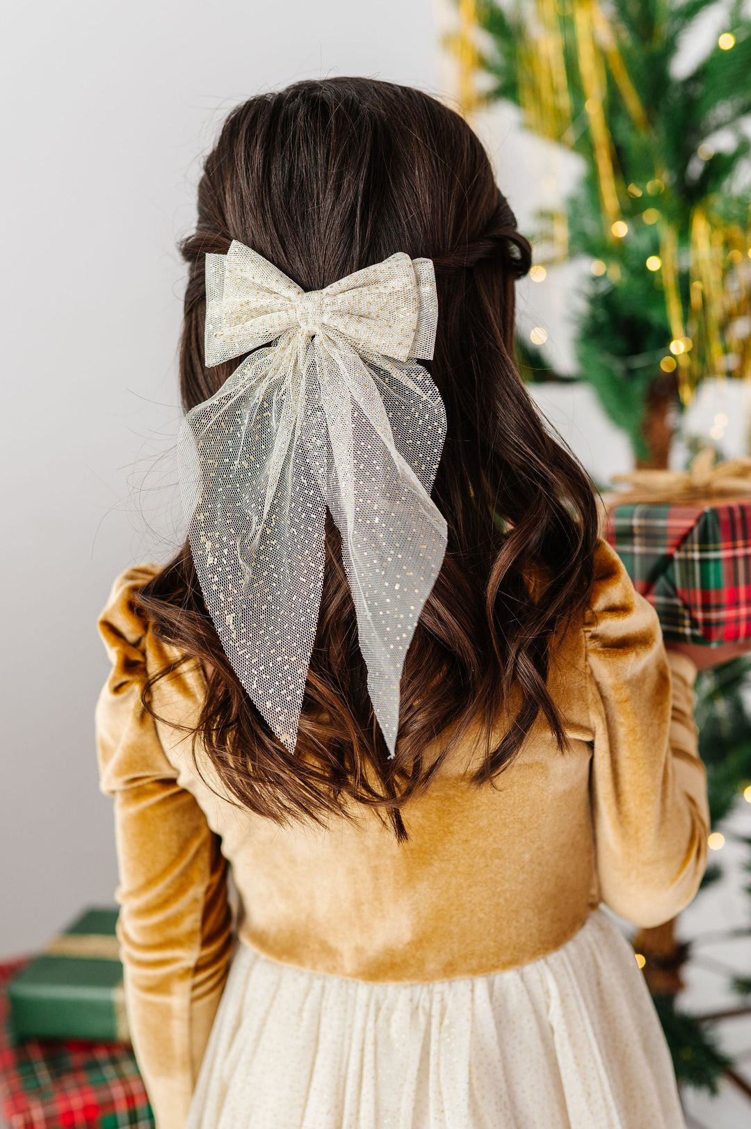 A child viewed from behind wearing a large glittery gold bow in their dark brown hair, dressed in a gold and cream christmas dress, with a decorated Christmas tree and wrapped gifts in the background.