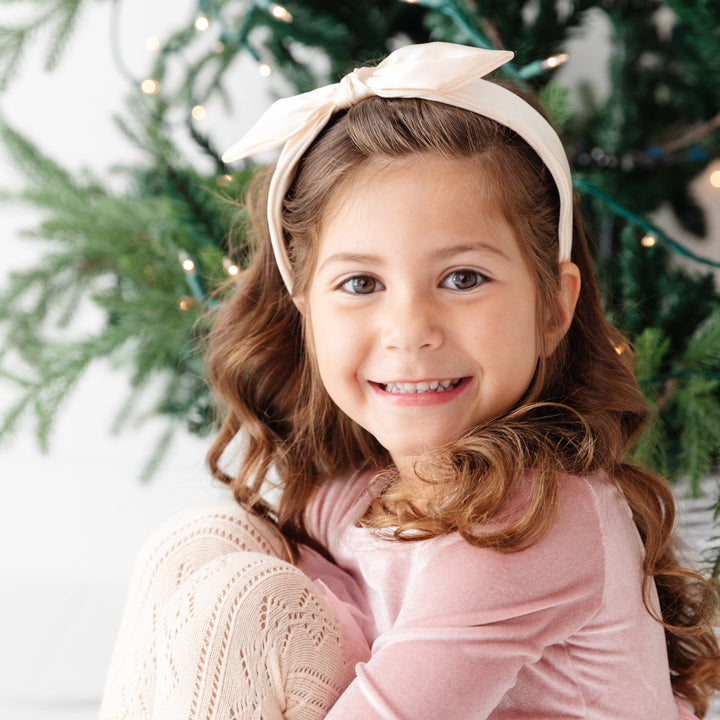 little girl in front of Christmas tree wearing cream colored satin headband and ballet pink velvet dress