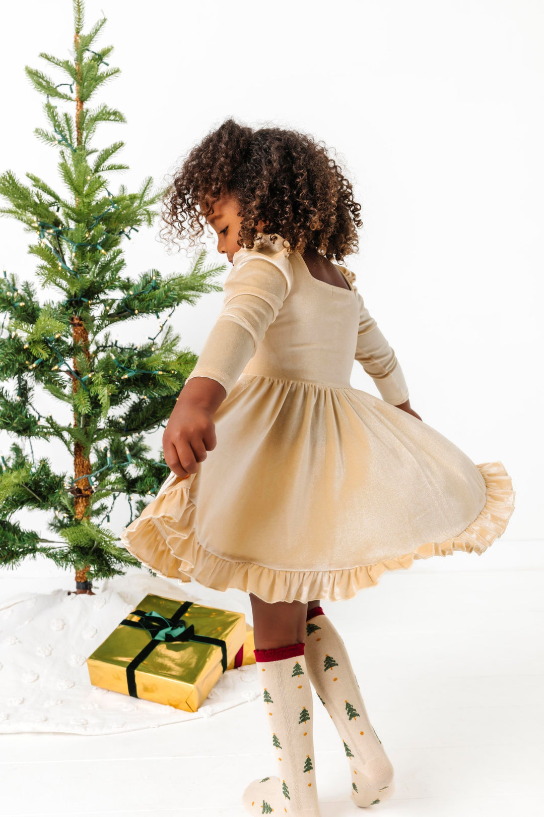 girl twirling in front of Christmas tree in cream velvet long sleeve twirl dress and Christmas tree print knee socks
