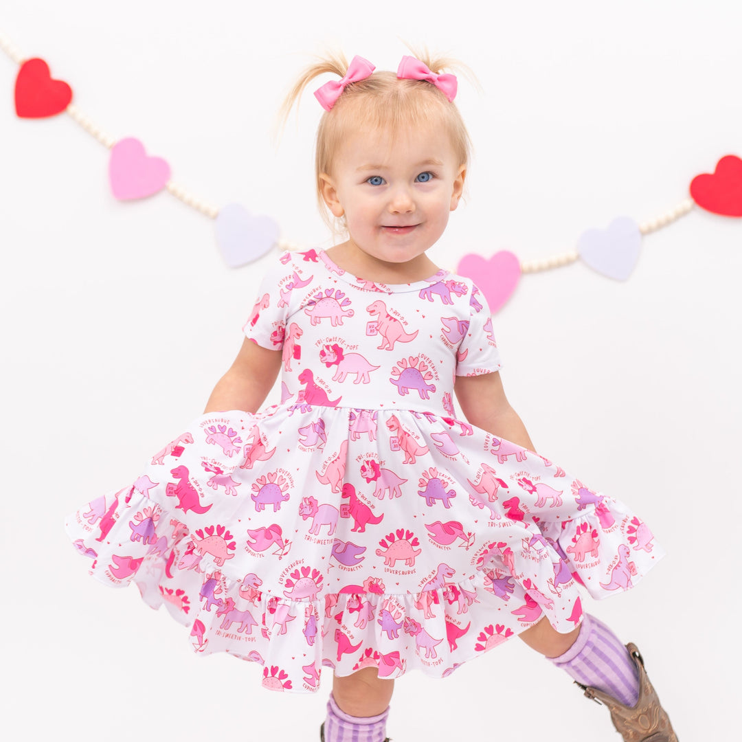 little girl with valentine dino print twirl dress in shades of pink and lavender with matching pink satin pigtail bows and lilac striped knee highs