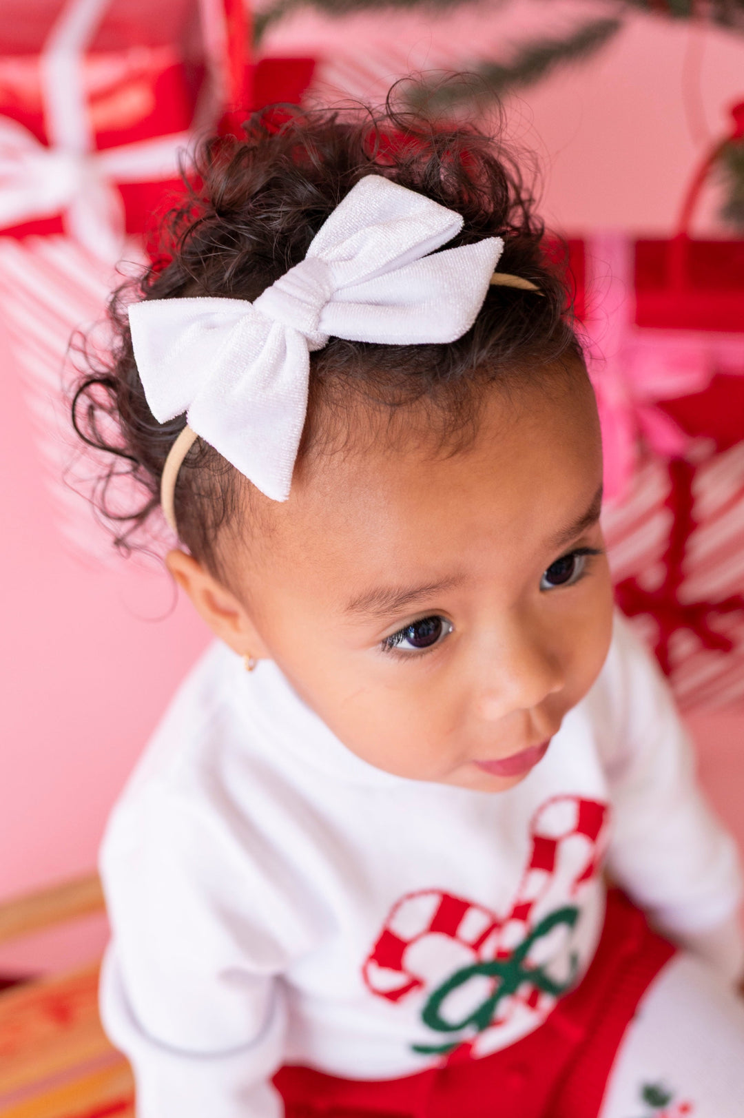 A toddler wearing a white bow headband, a white long-sleeve top with a red and green candy cane graphic, and red shorts, posed in front of a pink background with wrapped gifts.