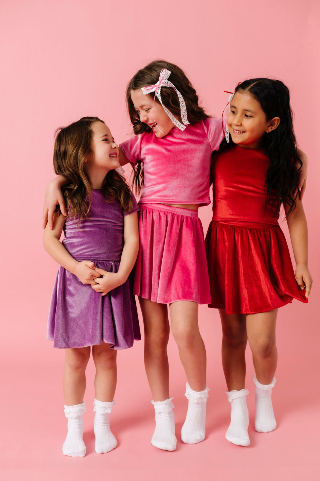 three girls in matching velvet top and skort sets in lavender, taffy pink and bright red paired with matching lace and ribbon hair bows and white ruffled bobby socks