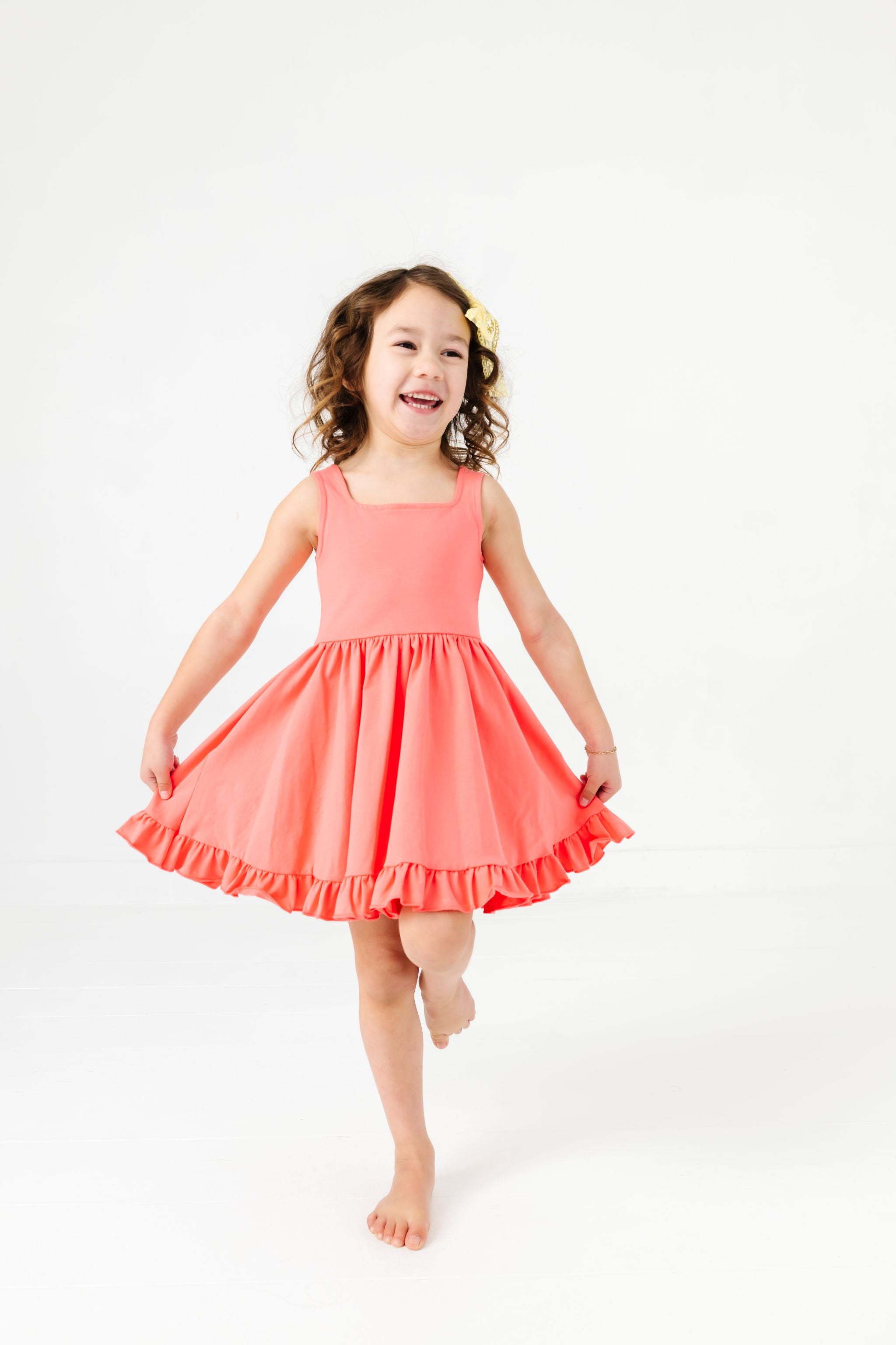 little girl in square neck sleeveless twirl dress with ruffle hem in shade of coral pink 