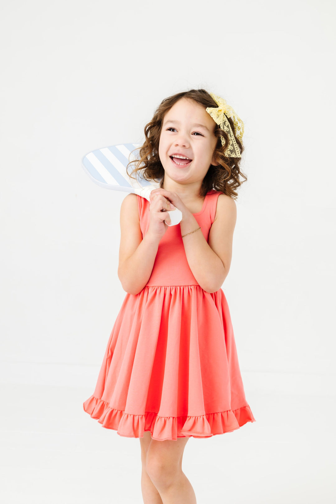 young girl holding pickleball racket in coral sleeveless twirl dress and yellow lace hair bow