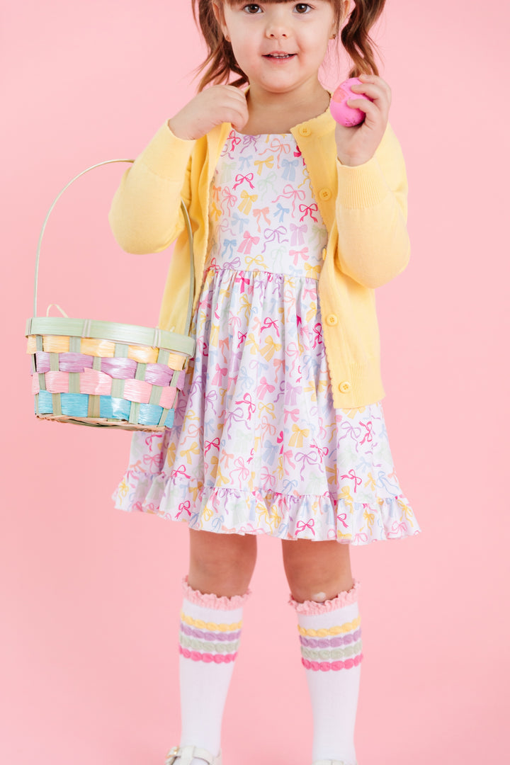 girl in easter outfit of square neck twirl dress in white with pastel multicolor bow print and matching knee highs and yellow cardigan