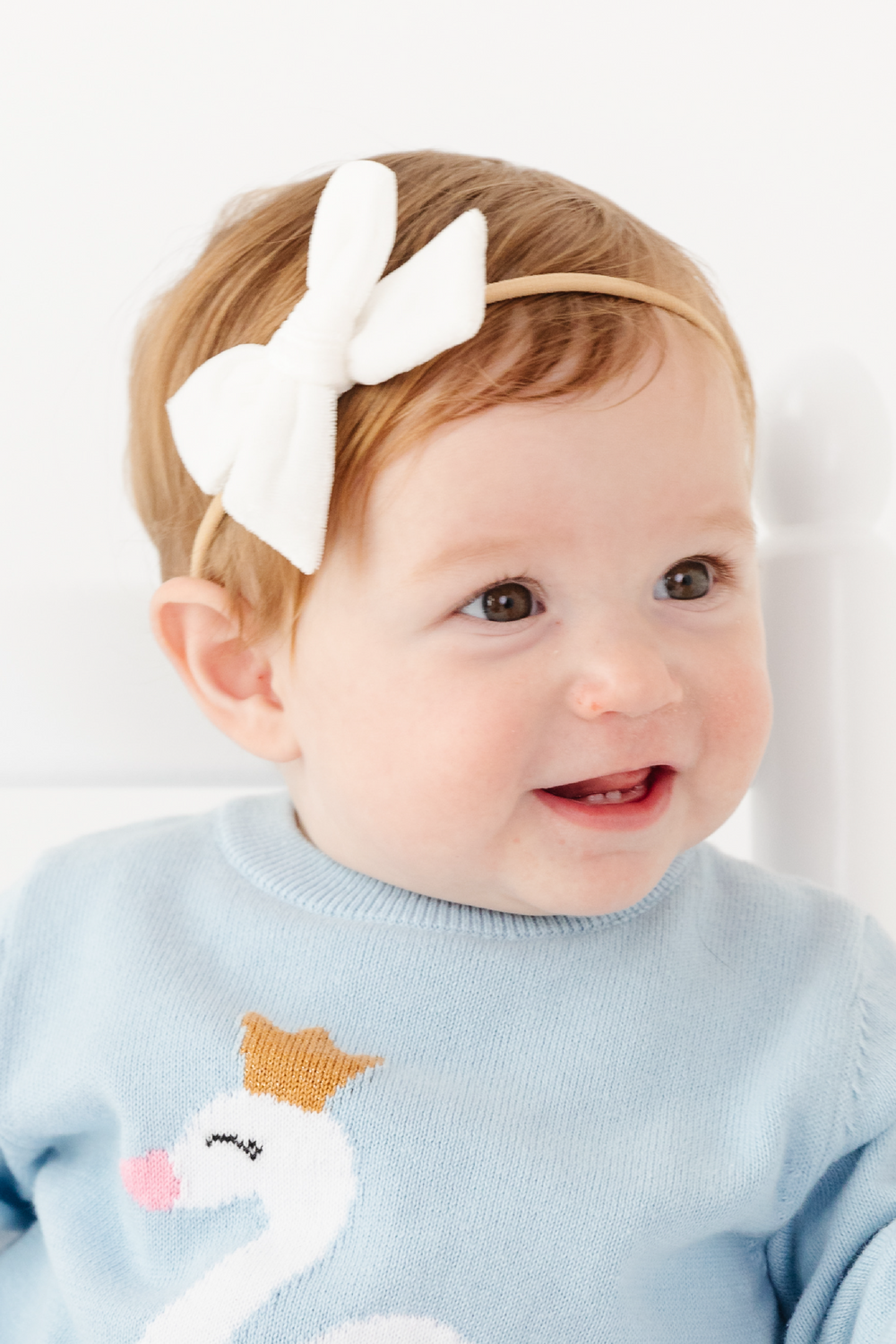 A baby wearing a white fabric bow headband on a beige elastic band, paired with a light blue knit sweater featuring a white swan design.