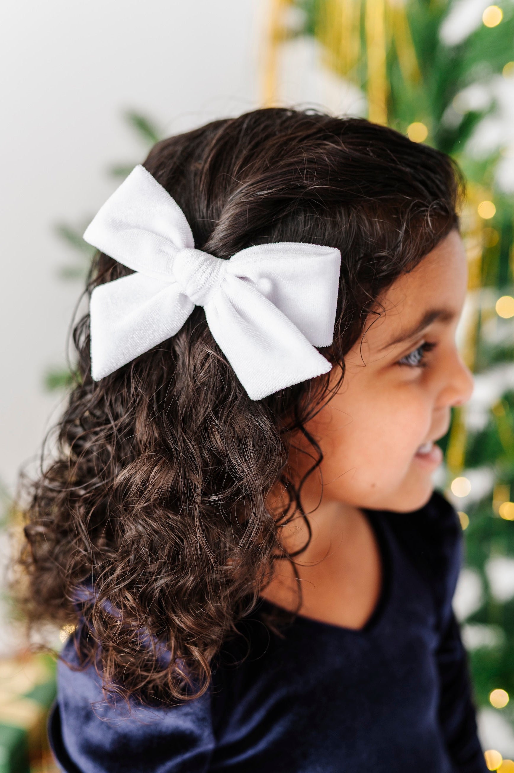 A young girl with dark curly hair wearing a large white fabric bow headband, shown in profile against a softly blurred Christmas tree background.