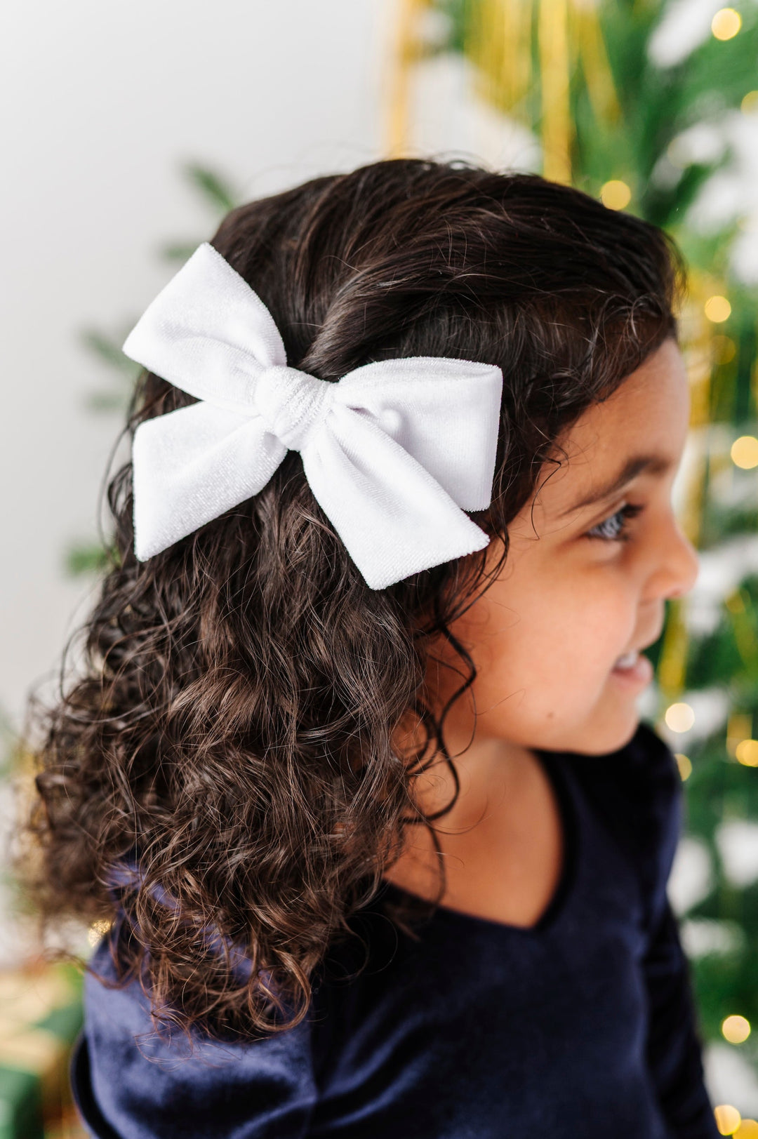 A young girl with dark curly hair wearing a large white fabric bow headband, shown in profile against a softly blurred Christmas tree background.