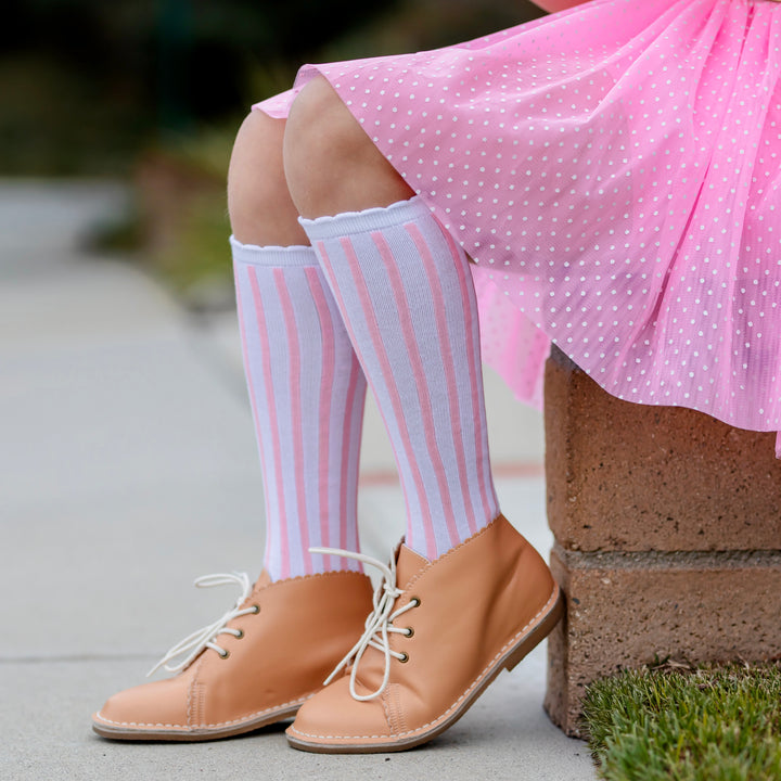 girl sitting with pastel pink tutu skort with white dots paired with white and pink vertical striped knee-high socks with white scalloped trim 