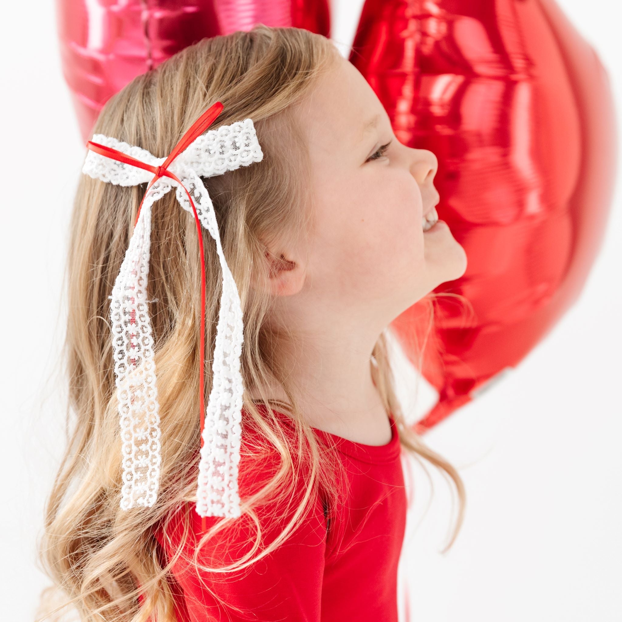 little girl holding heart balloons in long white lace bow with red ribbon detail and matching red valentine's dress