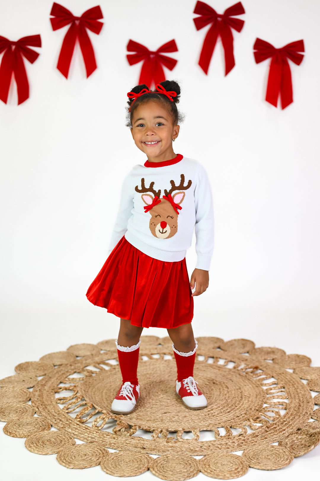 little girl in white knit Christmas sweater with Rudolph design and matching red velvet skirt, red knit pigtail bows, and red with white lace knee high socks