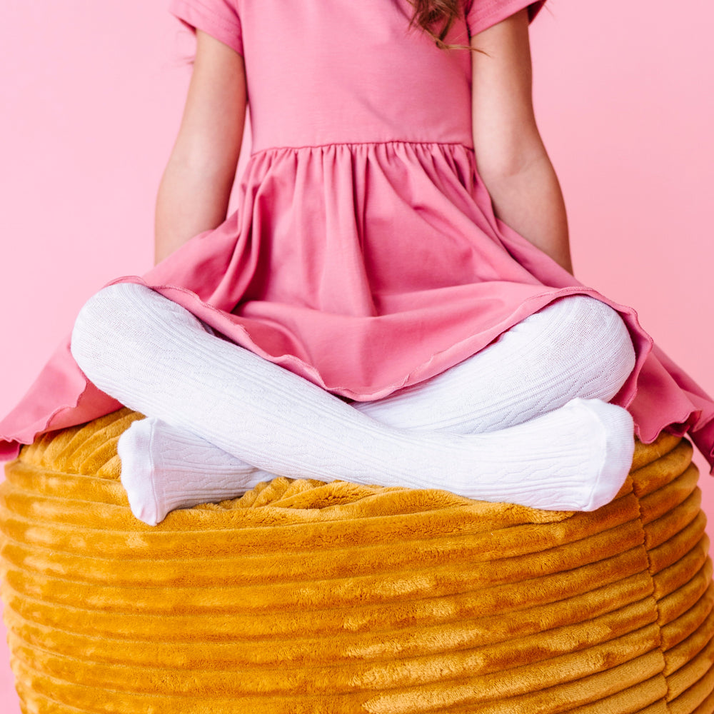 closeup of young girl sitting on mustard cushion in white cable knit tights paired with pink fall dress
