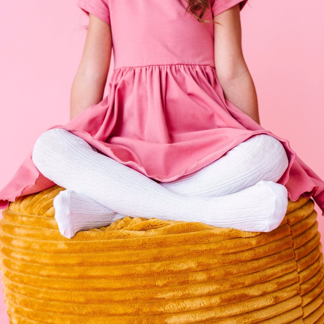 closeup of young girl sitting on mustard cushion in white cable knit tights paired with pink fall dress