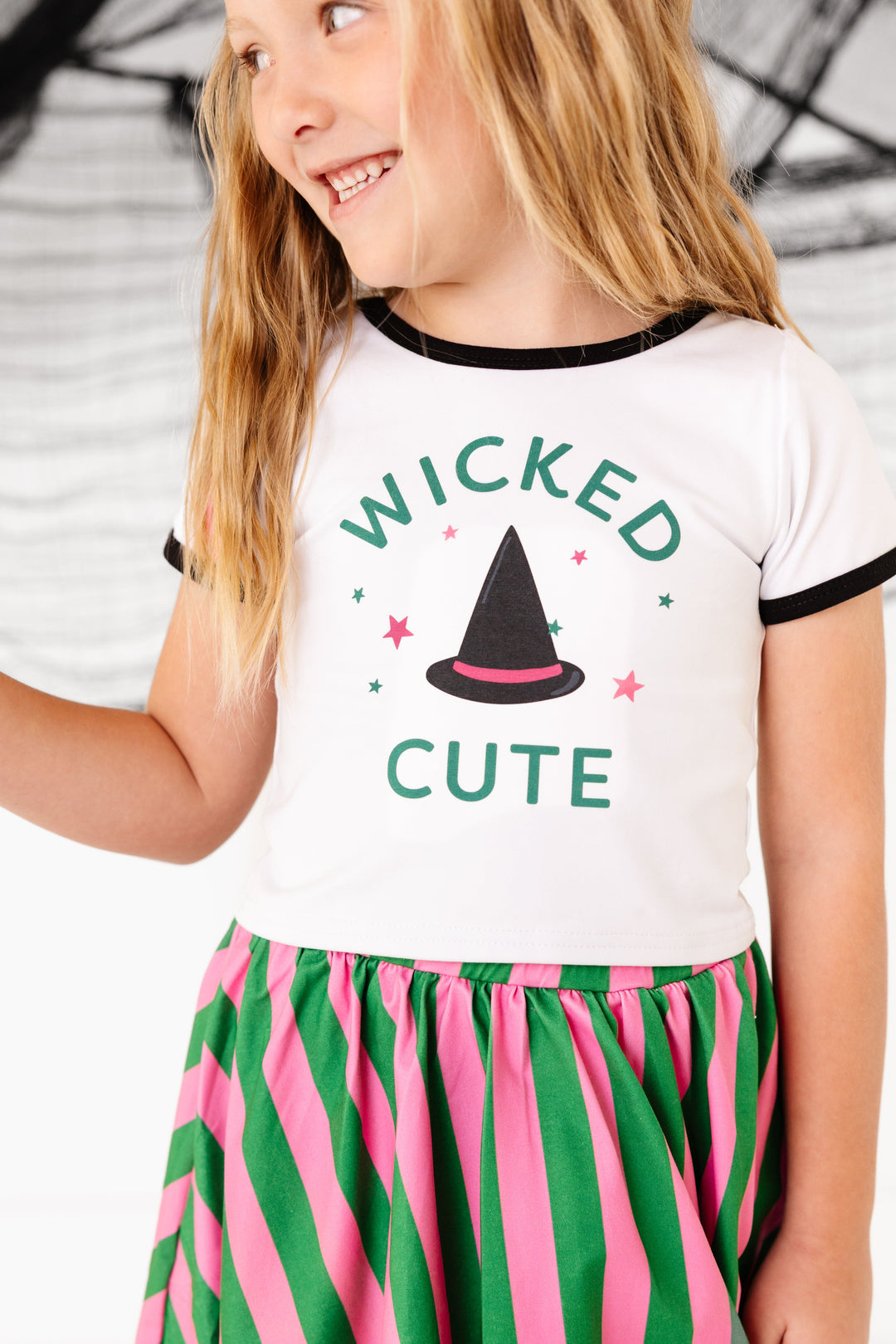 closeup of little girl in white with black binding short sleeve top with "Wicked Cute" phrase and witch hat design paired with green and pink striped skort
