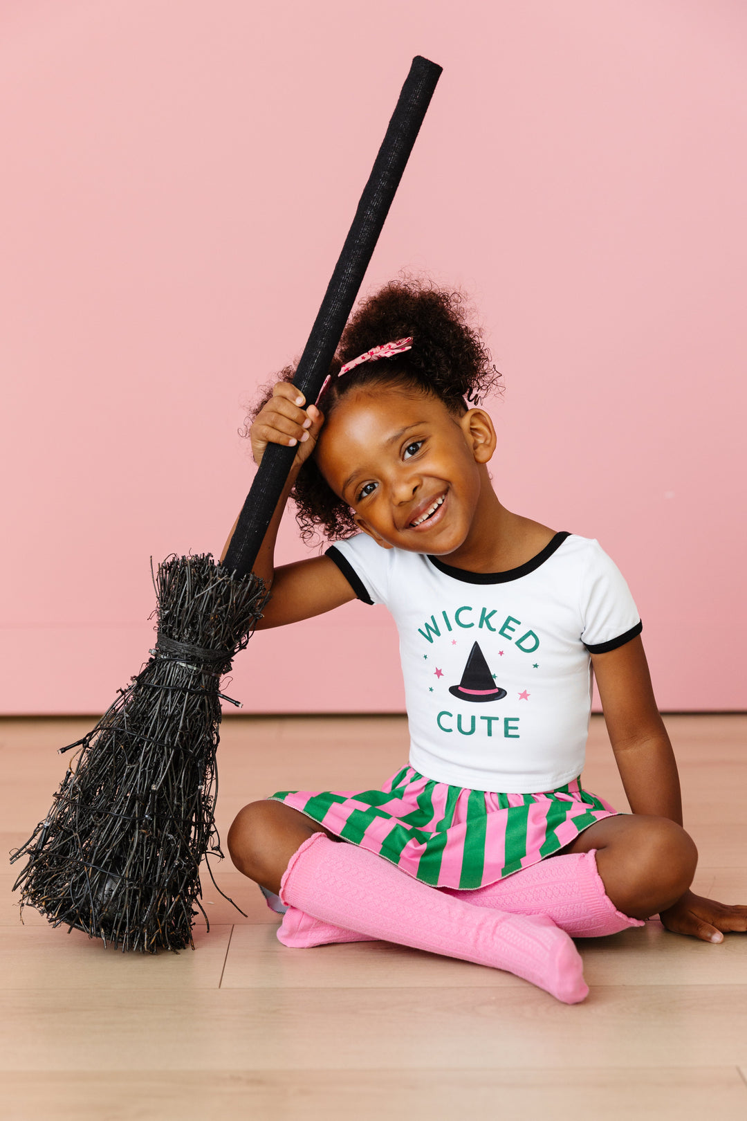 little girl holding broom in Wicked Cute witch hat tee paired with green and pink striped skort and matching pink socks and bows for Wicked inspired outfit 