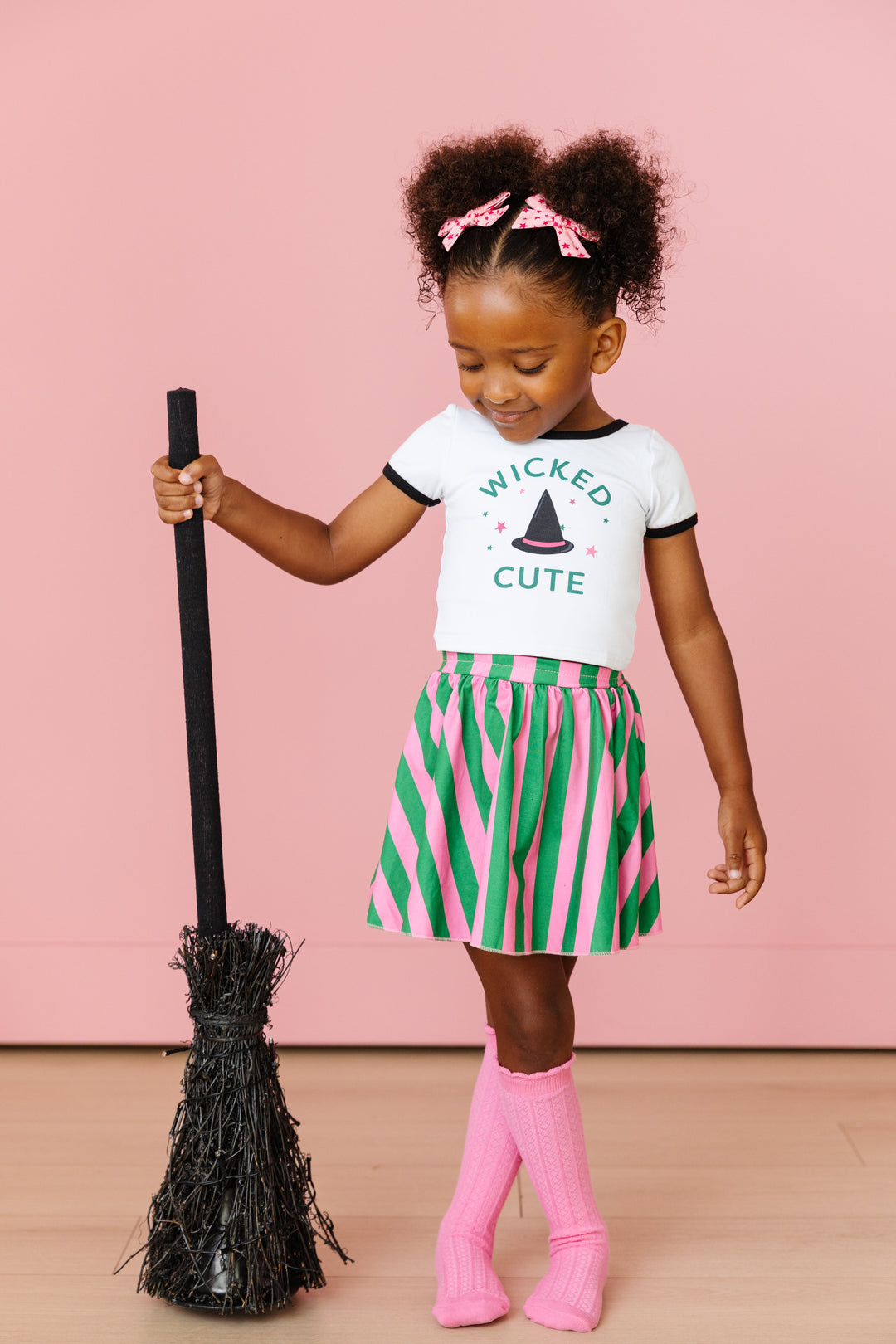 little girl holding broom in wicked cute witch top and matching green and pink striped skort with pink accesories