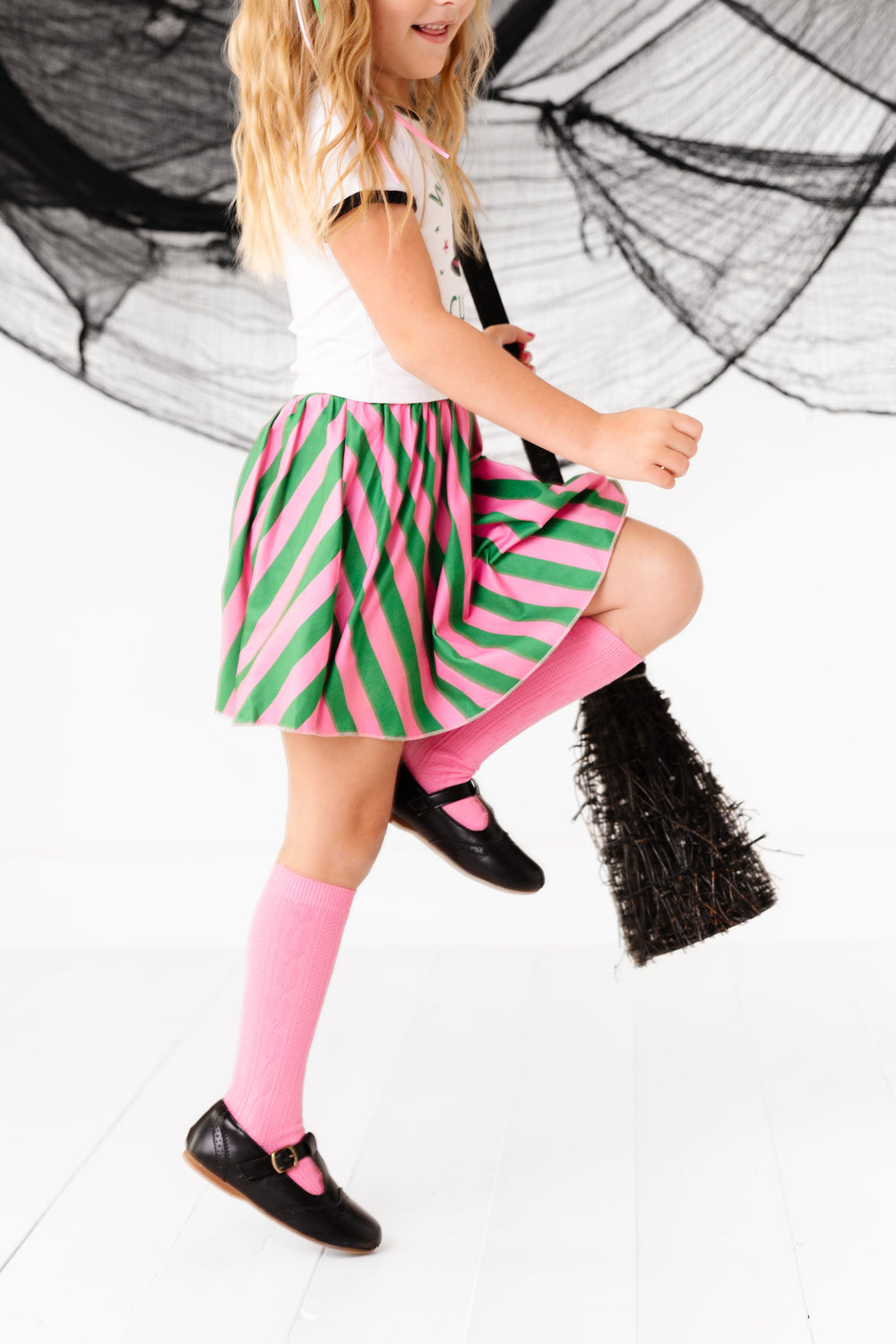 little girl jumping with broom in green and pink striped skort and matching pink cable knit knee highs for wicked inspired outfit