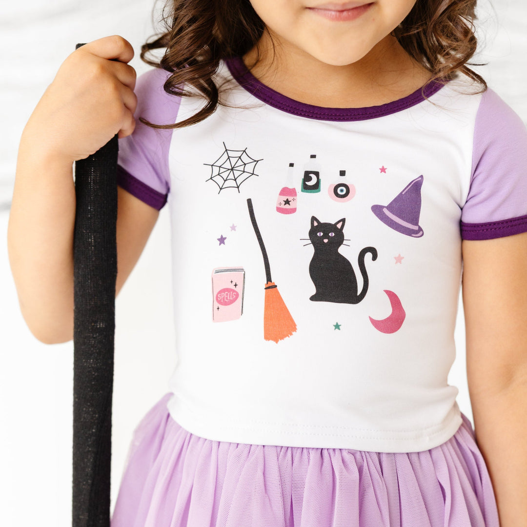 little girl in white shirt with purple sleeves and binding with witchy themed design and matching lavender tutu skort
