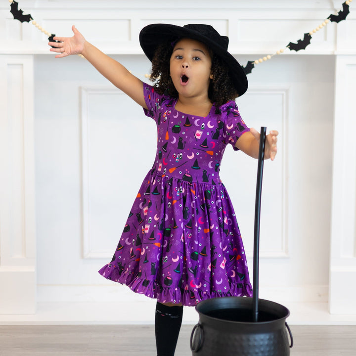 little girl with witch cauldron and hat wearing purple witchy print twirl dress and matching black cat knee high socks