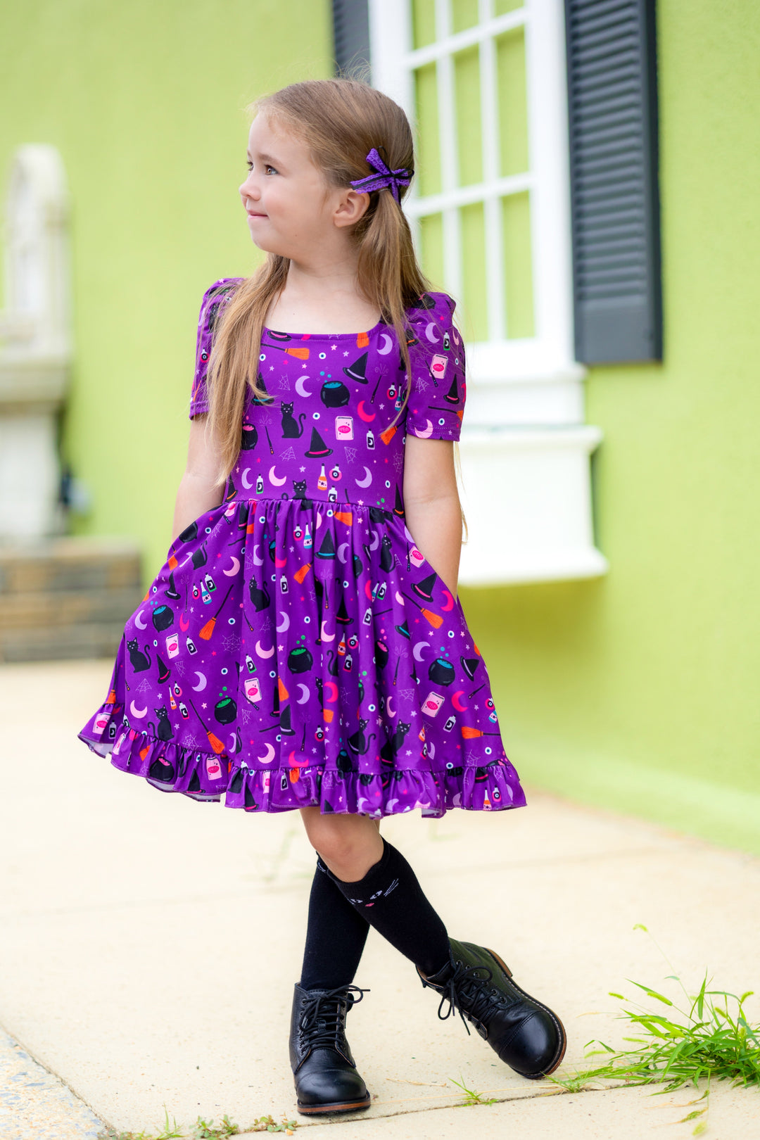 little girl wearing purple Halloween twirl dress with witchy vibe print, matching black cat knee high socks and purple lace pigtail bows