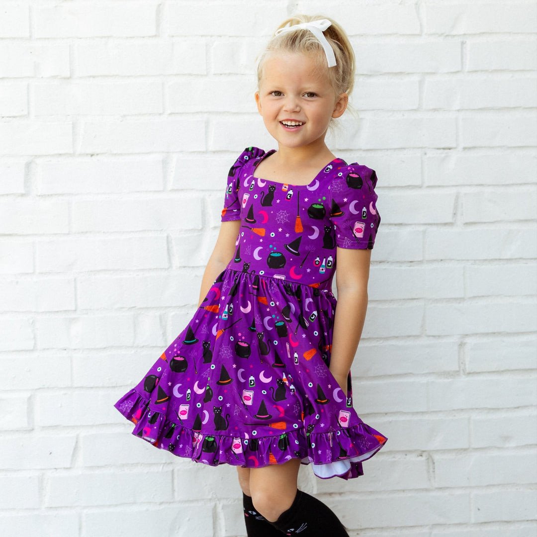 Young girl in a purple Halloween dress standing against a white brick wall with decorative bats.