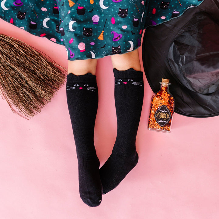 girl sitting on pink backdrop in teal witchy print dress paired with black knee socks with cat ears and cat face design