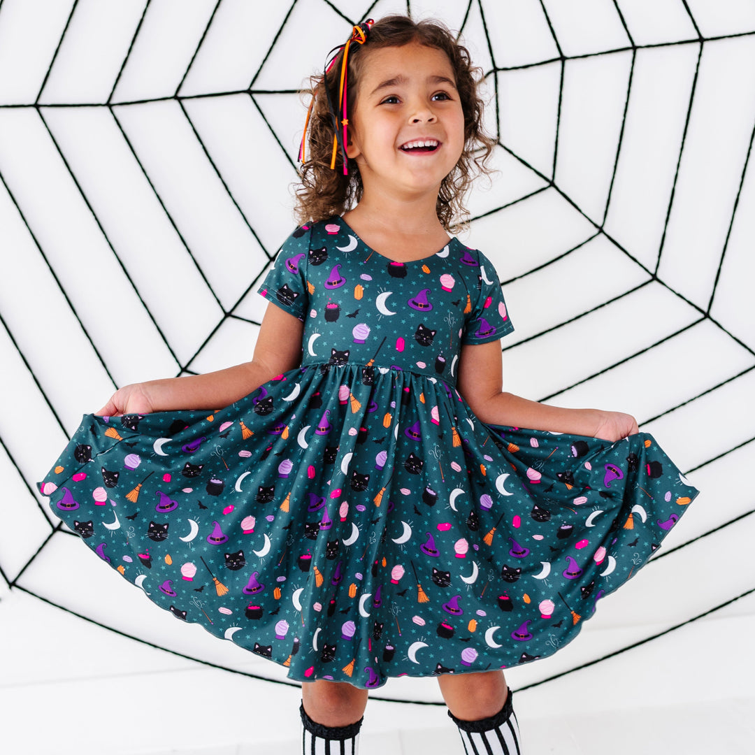young girl in front of faux spider web wearing team witch themed printed v-neck dress paired with star ribbon hair bow and black and white striped lace top knee socks