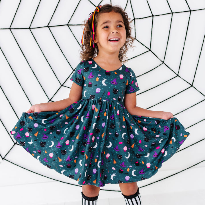 young girl in front of faux spider web wearing team witch themed printed v-neck dress paired with star ribbon hair bow and black and white striped lace top knee socks