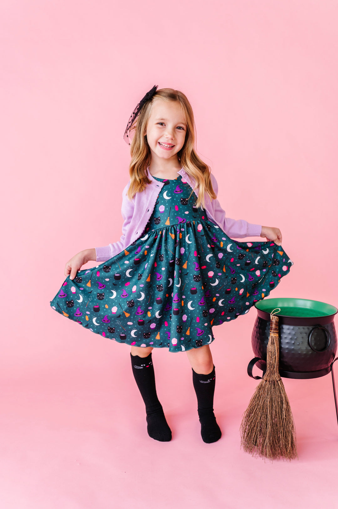 young girl by cauldres in teal witchy themed twirl dress paired with lavender cotton knit cardigan and black cat knee socks