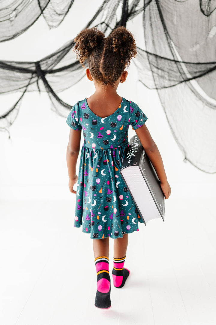 little girl walking away from camera holding large spell book to show v-neck design on teal witchy themed twirl dress paired with black, pink and orange striped midi socks