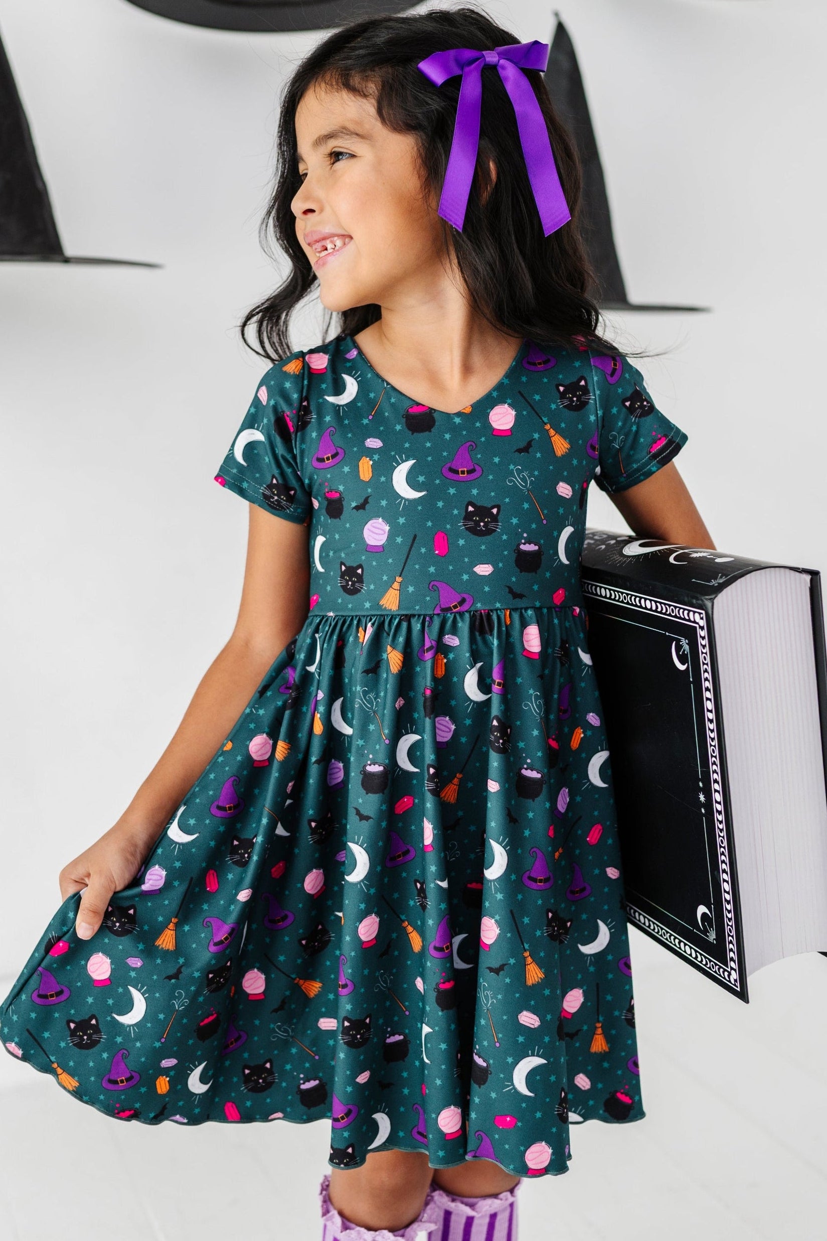 young girl holding large spell book in teal v-neck twirl dress with cute, classic witch themed print paired with purple accessories for Halloween