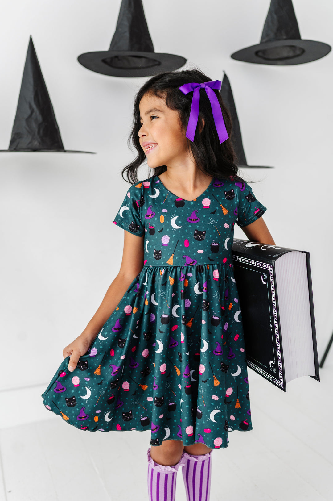 young girl holding large spell book in teal v-neck twirl dress with cute, classic witch themed print paired with purple accessories for Halloween