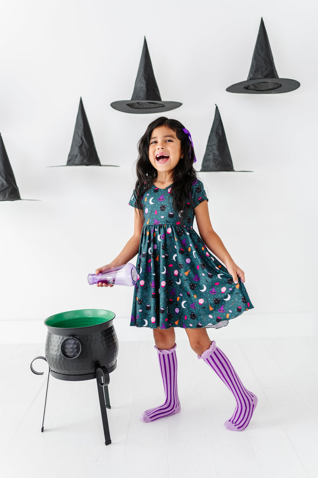 little girl surrounded by floating witch hats while pouring potion into cauldron wearing teal witchy themed twirl dress paired with two-tone purple striped knee socks